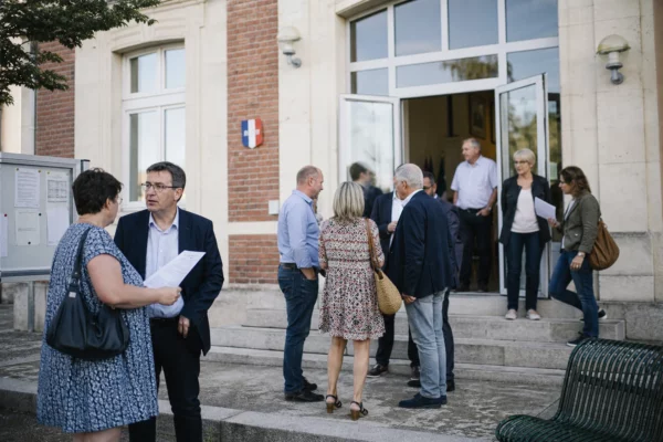 Des habitants échangent devant une mairie de province, dans une ambiance politique locale claire et naturelle.