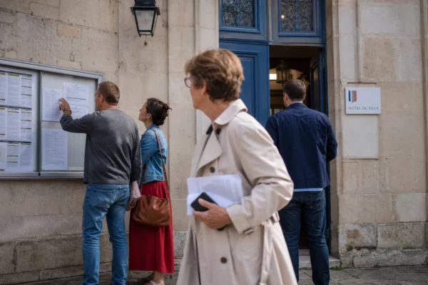 Des passants anonymes discutent devant une mairie française, près d’un panneau d’information public.