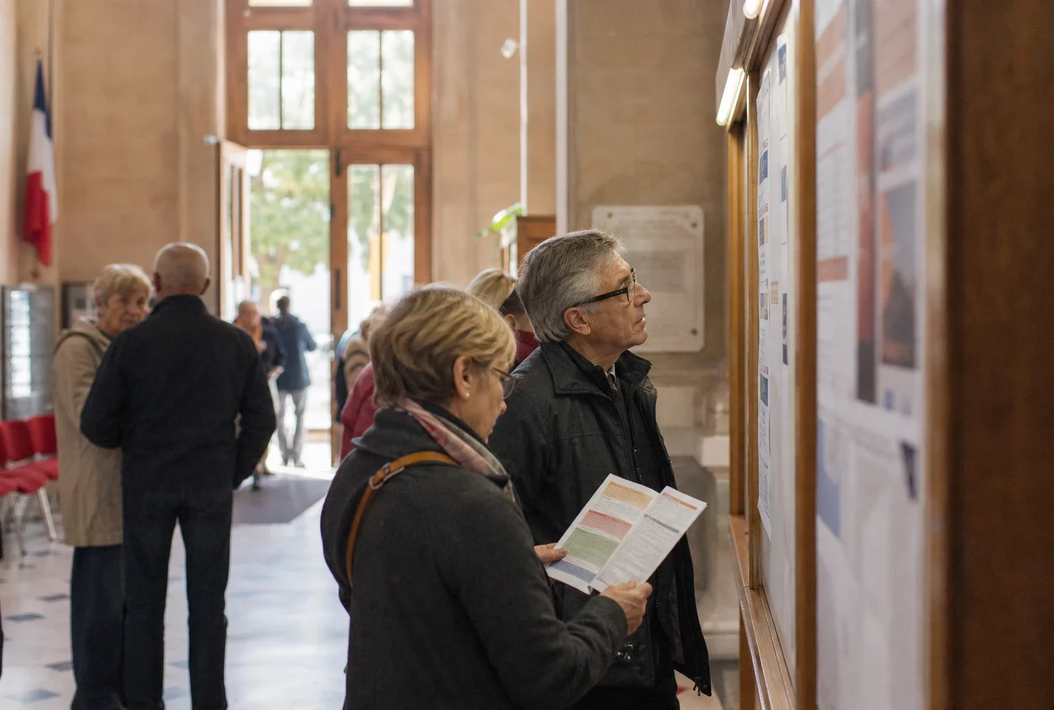 Hall de mairie français avec des habitants anonymes devant un panneau d’affichage flou et une lumière naturelle claire.