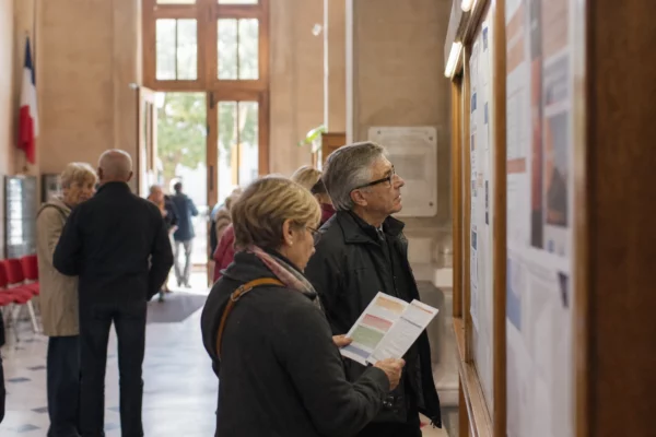 Hall de mairie français avec des habitants anonymes devant un panneau d’affichage flou et une lumière naturelle claire.