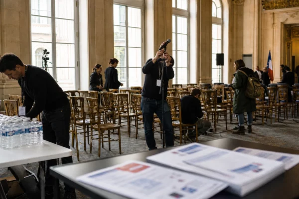 Salle de meeting parisienne en préparation, avec chaises alignées, micros de presse et staff anonyme sous la lumière du jour.