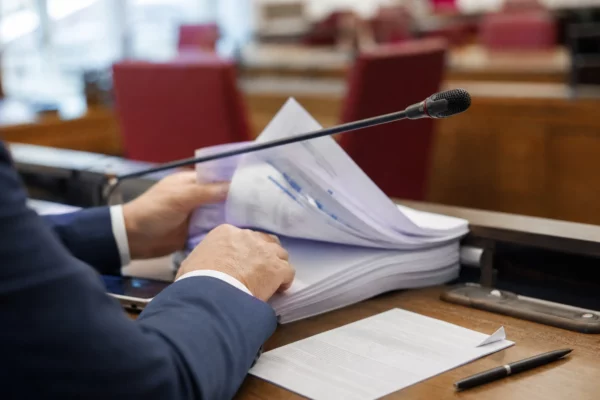 Maines anonymes feuilletant un dossier de commission avec micro de table dans un intérieur parlementaire lumineux