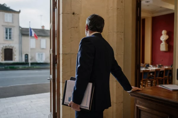 Un élu local anonyme dans une mairie, carnet à la main, sur fond de rue calme et de salle municipale.