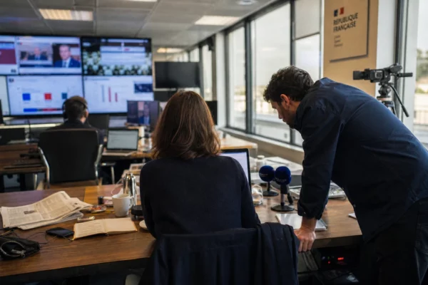 Dans une rédaction française, un journaliste prépare un sujet politique avec écrans flous, micros et documents anonymes.