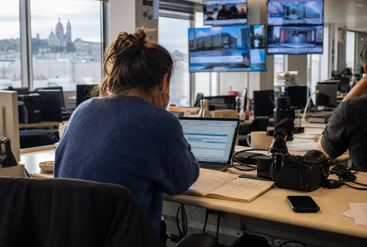 Dans une rédaction française, un journaliste prépare un sujet sur le parquet européen, avec écrans flous et matériel de presse.