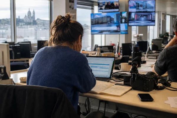 Dans une rédaction française, un journaliste prépare un sujet sur le parquet européen, avec écrans flous et matériel de presse.