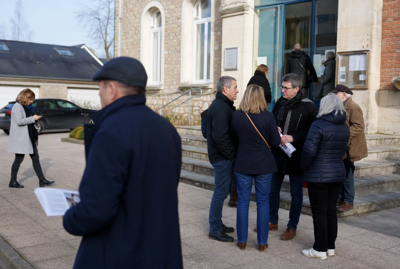 Des habitants anonymes arrivent devant une mairie lors d’une réunion locale, dans une ambiance claire et ordinaire.