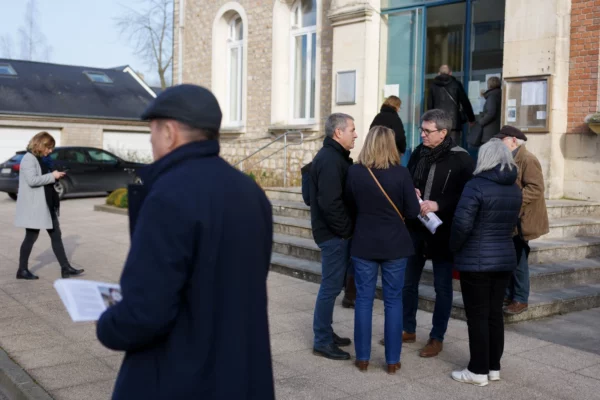 Des habitants anonymes arrivent devant une mairie lors d’une réunion locale, dans une ambiance claire et ordinaire.