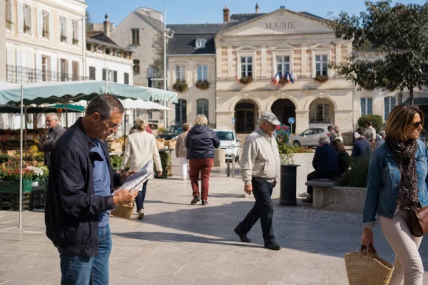 Scène de rue française près d’une mairie de ville moyenne, avec habitants anonymes et marché local en arrière-plan.