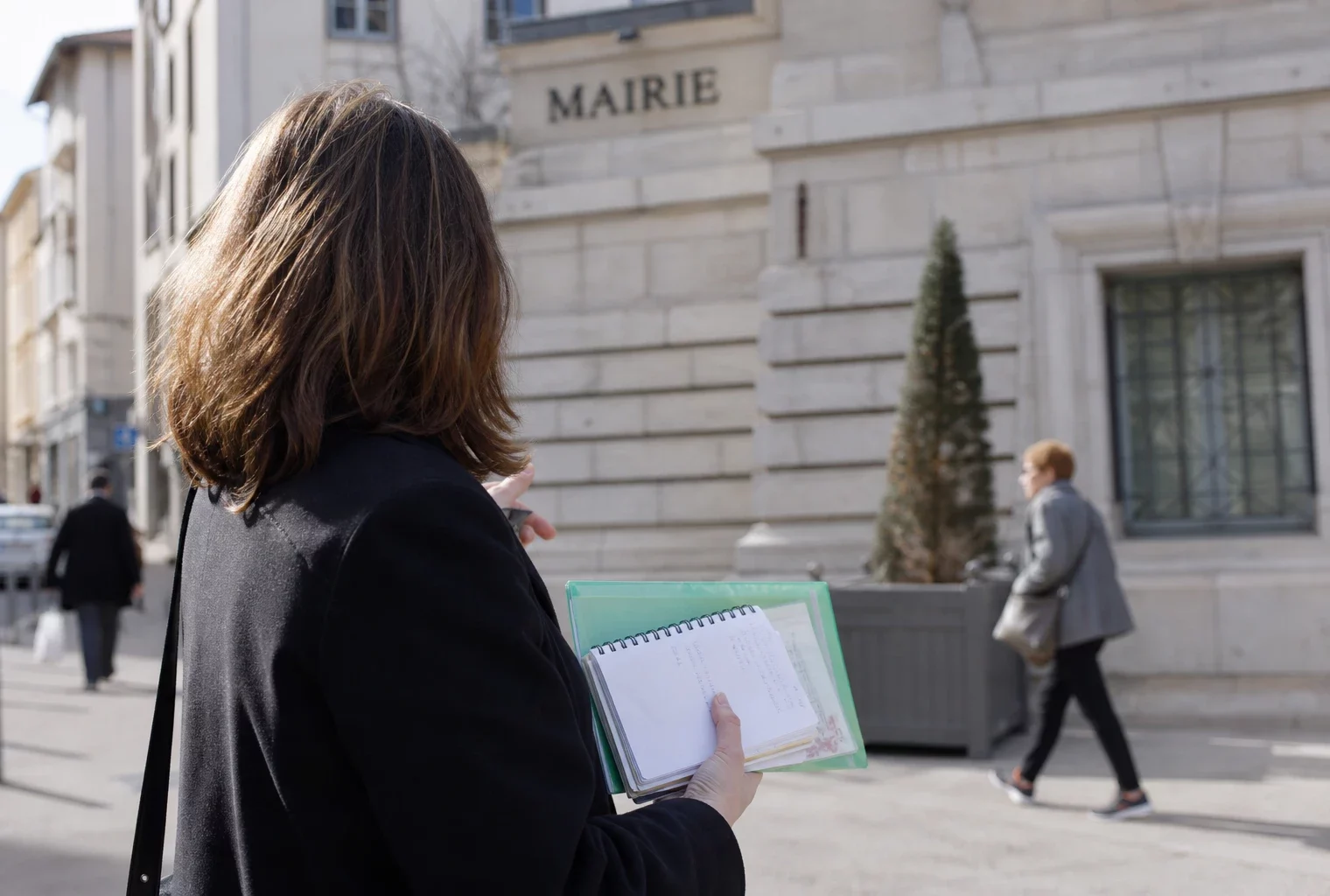 Collaborateur de campagne anonyme devant une mairie de Lyon, carnet en main, dans une lumière naturelle de rue.