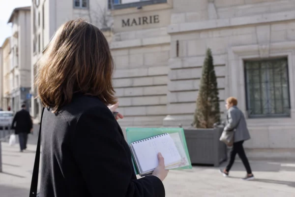 Collaborateur de campagne anonyme devant une mairie de Lyon, carnet en main, dans une lumière naturelle de rue.