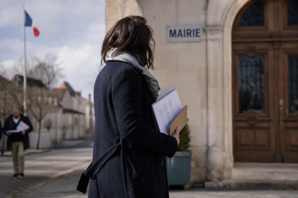 Portrait d’un élu local anonyme devant une mairie française, carnet en main, dans une lumière naturelle douce.