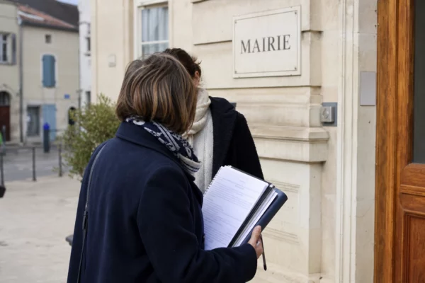 Un élu local anonyme note sur un carnet devant une mairie française, dans une scène de reportage claire et naturelle.