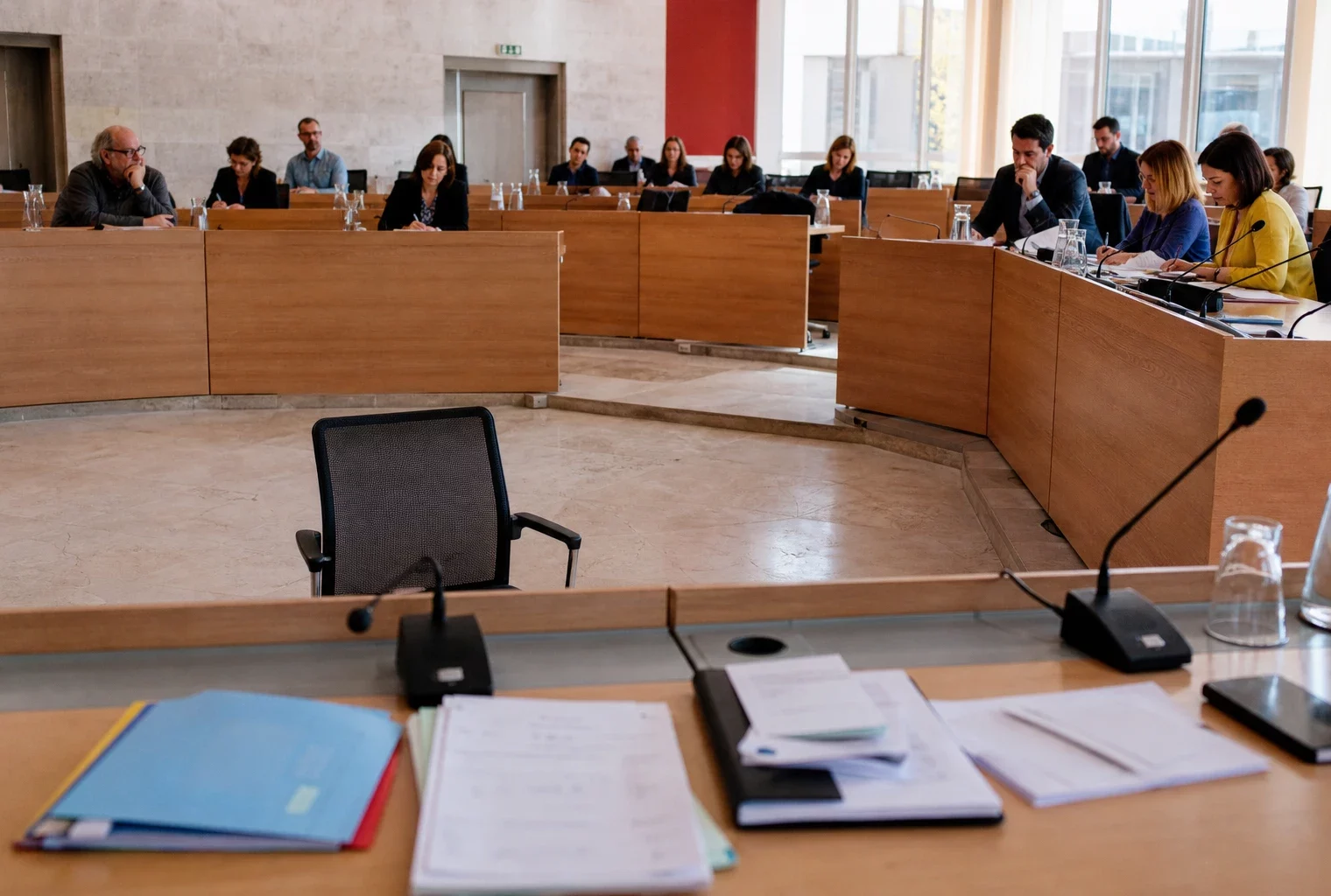 Salle municipale française lumineuse avec chaise vide, micros et dossiers flous, ambiance de réunion politique locale
