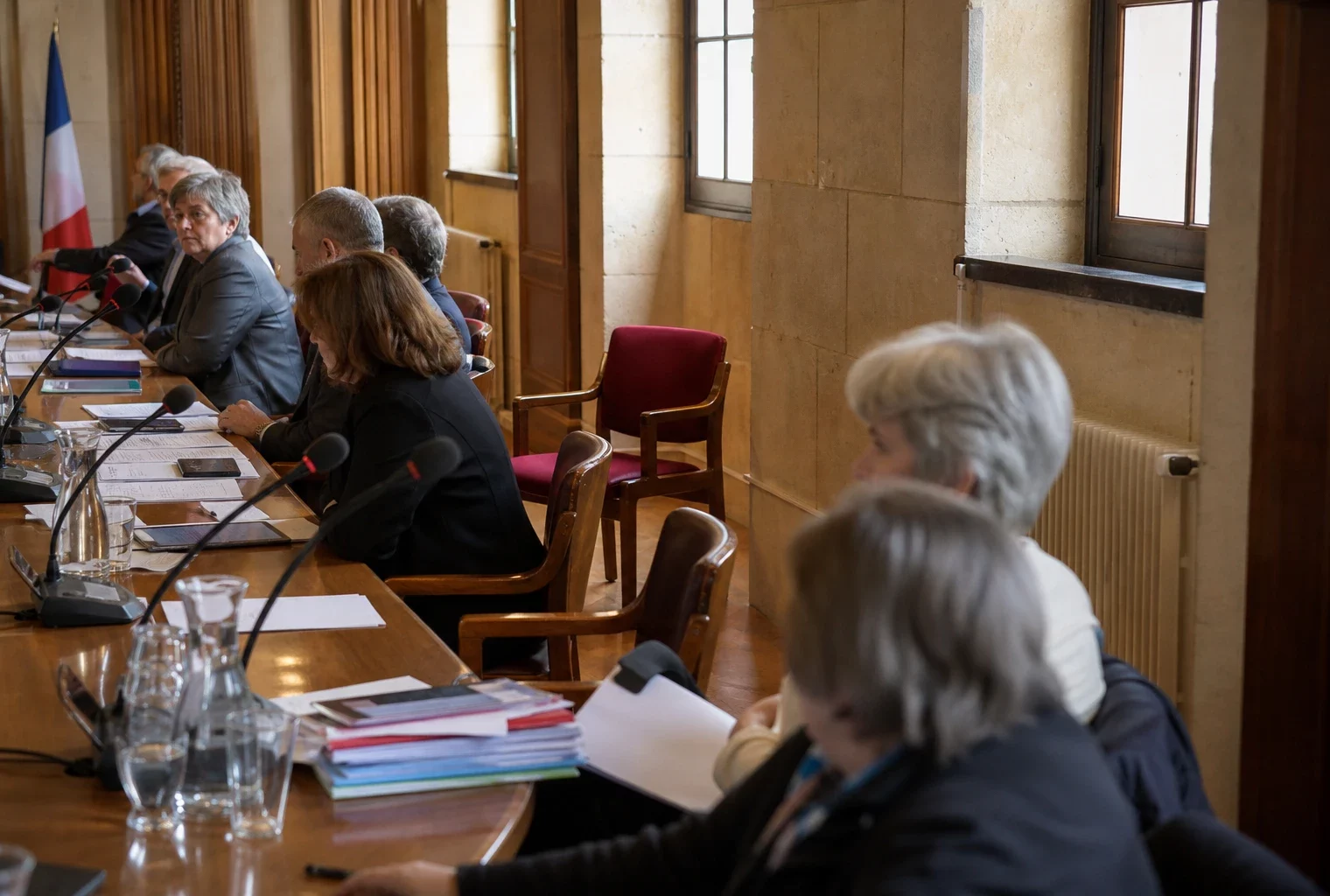 Salle municipale française avec micros, dossiers et une chaise vide lors d’une réunion politique locale