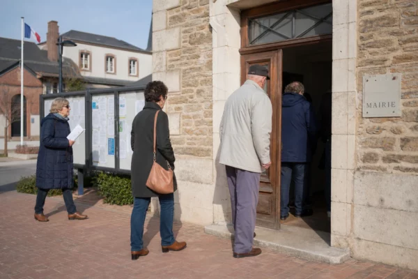Devant une mairie française, des citoyens anonymes assistent à une réunion locale sur fond de lumière naturelle.
