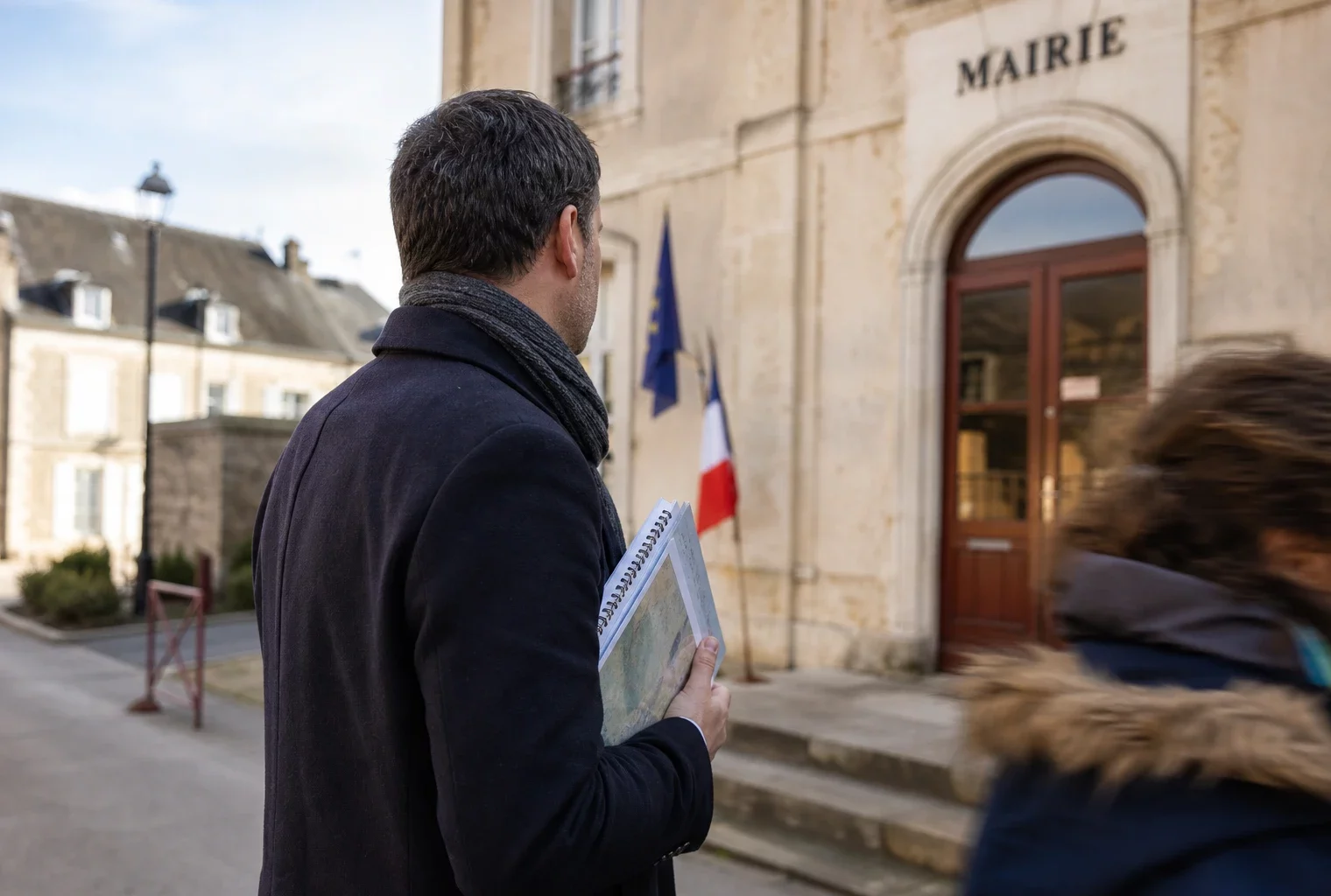 Un élu local anonyme de dos tient un dossier devant une mairie de province, dans une lumière naturelle et calme.