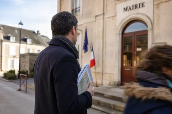 Un élu local anonyme de dos tient un dossier devant une mairie de province, dans une lumière naturelle et calme.