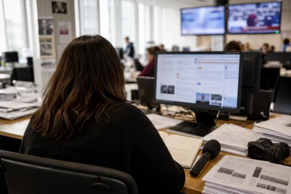 Journaliste de dos dans une rédaction française, entouré d’écrans flous et de documents de travail