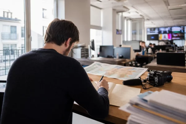 Journaliste en rédaction préparant une analyse politique, carnet ouvert, micro sans logo et écrans flous en arrière-plan.