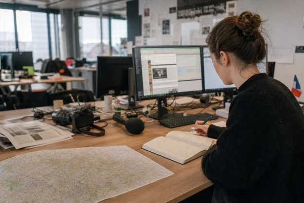 Journaliste dans une rédaction française préparant un sujet politique avec carnet, micro sans logo et carte floue.