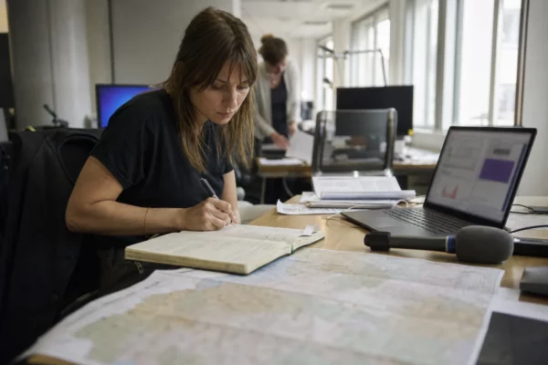 Journaliste en rédaction préparant un sujet territorial sur la canicule, avec carnet, micro et carte floue.