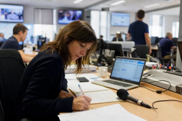 Journaliste en rédaction préparant un sujet sur le Défenseur des droits, avec micro, carnet et écrans flous.
