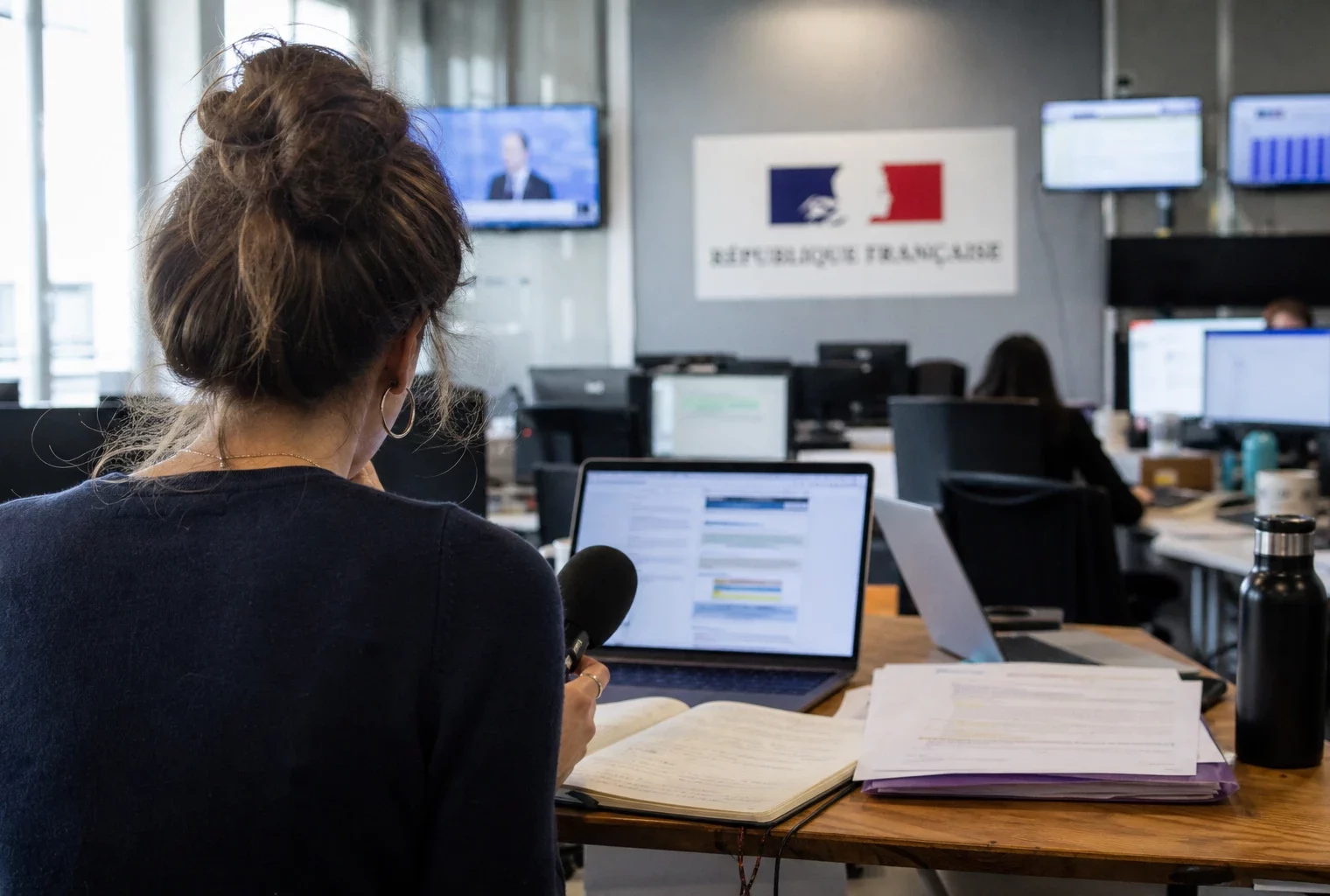 Journaliste dans une rédaction française préparant un sujet sur Sciences Po et ESCP, avec micro, carnet et écrans flous.