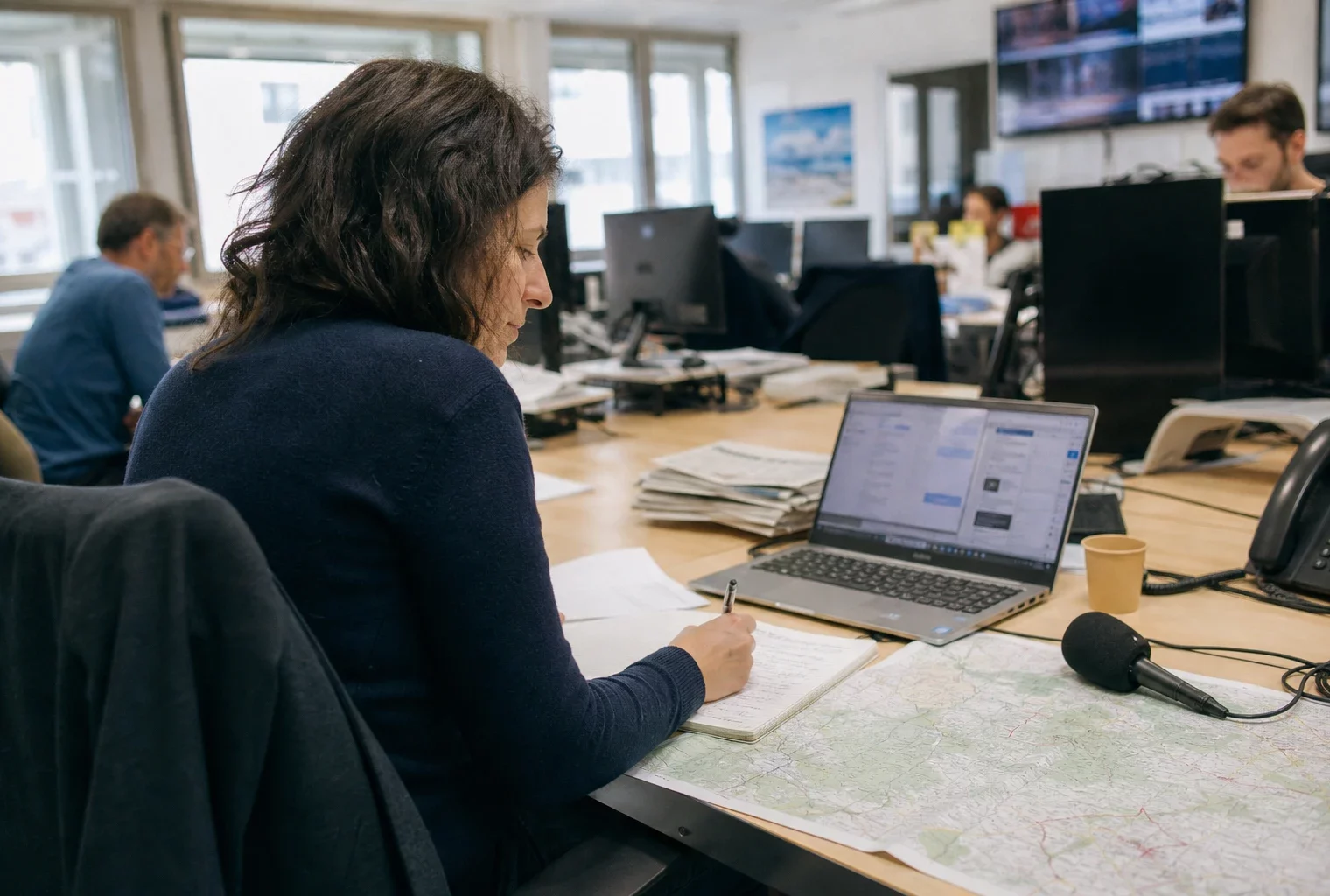 Journaliste dans une rédaction française, carnet ouvert et micro sans logo, avec écrans flous en arrière-plan.