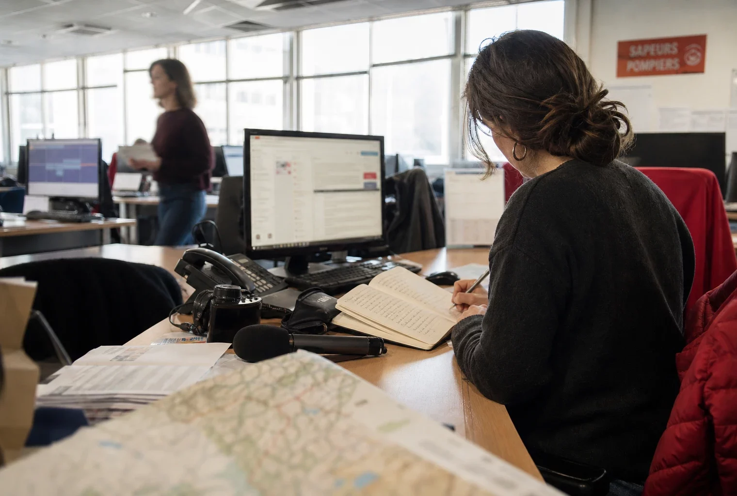 Journaliste en rédaction préparant un sujet territorial sur les incendies, avec carnet, micro et carte floue.