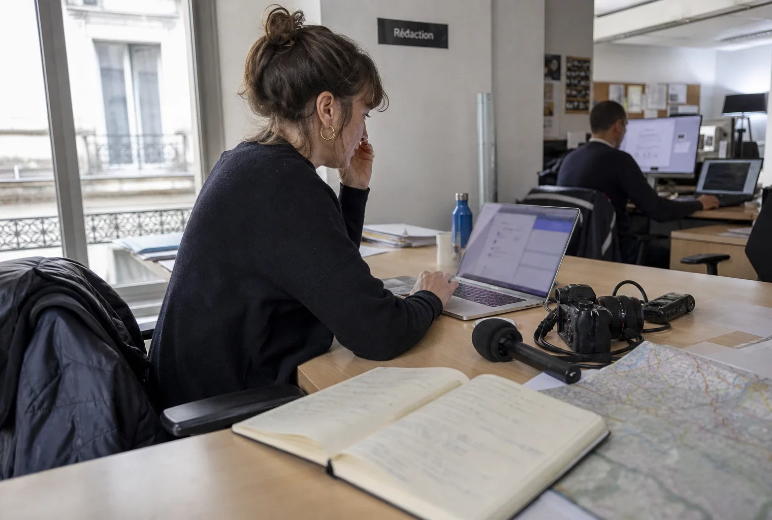 Journaliste en rédaction préparant un sujet territorial avec carnet, micro et carte floue près d’une fenêtre lumineuse.