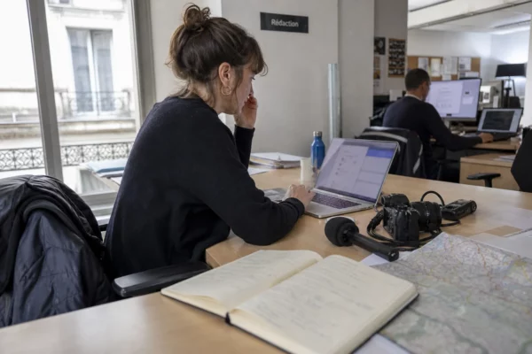 Journaliste en rédaction préparant un sujet territorial avec carnet, micro et carte floue près d’une fenêtre lumineuse.