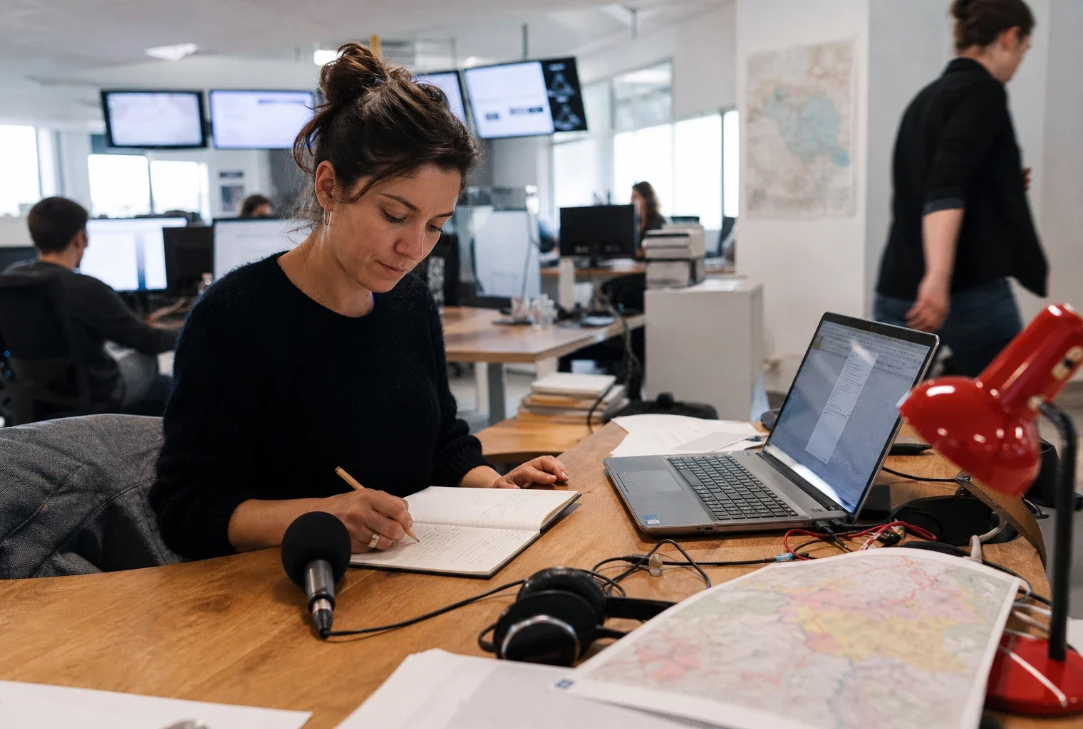 Journaliste en rédaction préparant un sujet sur les incendies et le Tour de France, avec carnet, carte floue et micro sans logo.