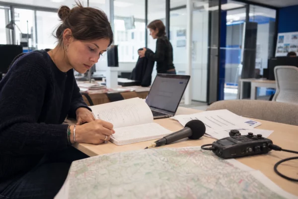 Journaliste dans une rédaction française préparant un sujet sur les hôpitaux et la canicule, carnet, micro et carte floue.