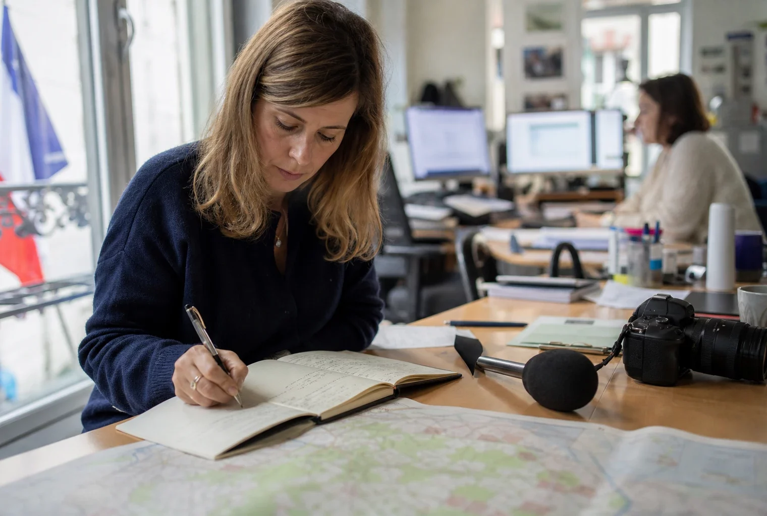Journaliste en rédaction préparant un sujet territorial avec carnet, micro sans logo et carte papier floue.