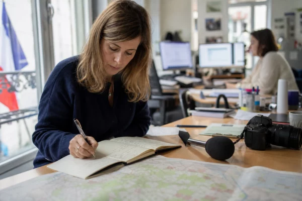 Journaliste en rédaction préparant un sujet territorial avec carnet, micro sans logo et carte papier floue.