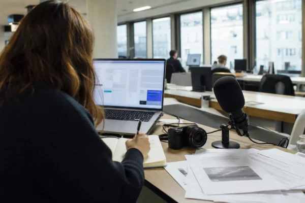 Journaliste en rédaction préparant un sujet de justice, avec micro, carnet et écran d’ordinateur flou.
