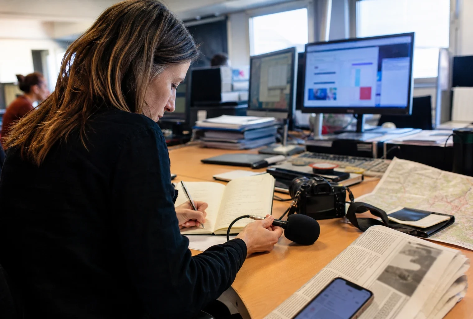 Journaliste en rédaction préparant un sujet sur les cagnottes liées aux incendies, avec carnet, micro et écrans flous.