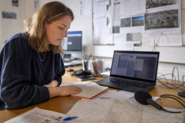 Journaliste dans une rédaction locale préparant un sujet sur Grenoble, avec carnet, micro sans logo et carte papier floue.