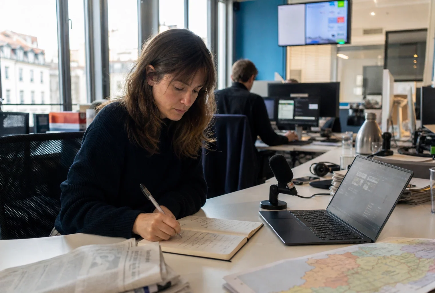 Journaliste dans une rédaction locale préparant un sujet sur l’IA, avec carnet, micro et écran flou.