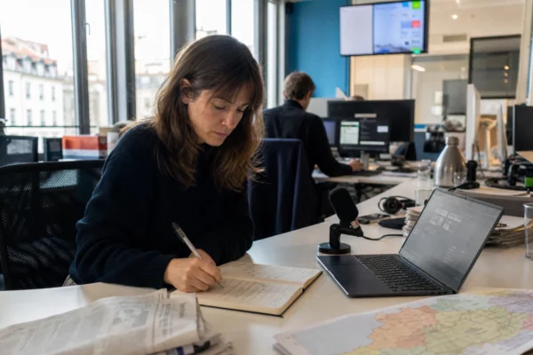 Journaliste dans une rédaction locale préparant un sujet sur l’IA, avec carnet, micro et écran flou.