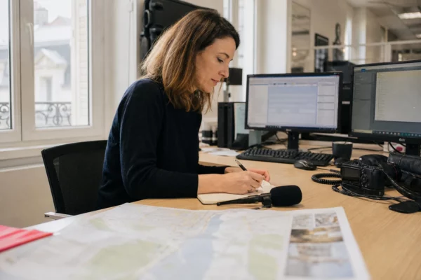 Journaliste en rédaction préparant un sujet local avec carnet, micro sans logo et carte papier floue près d’une fenêtre.