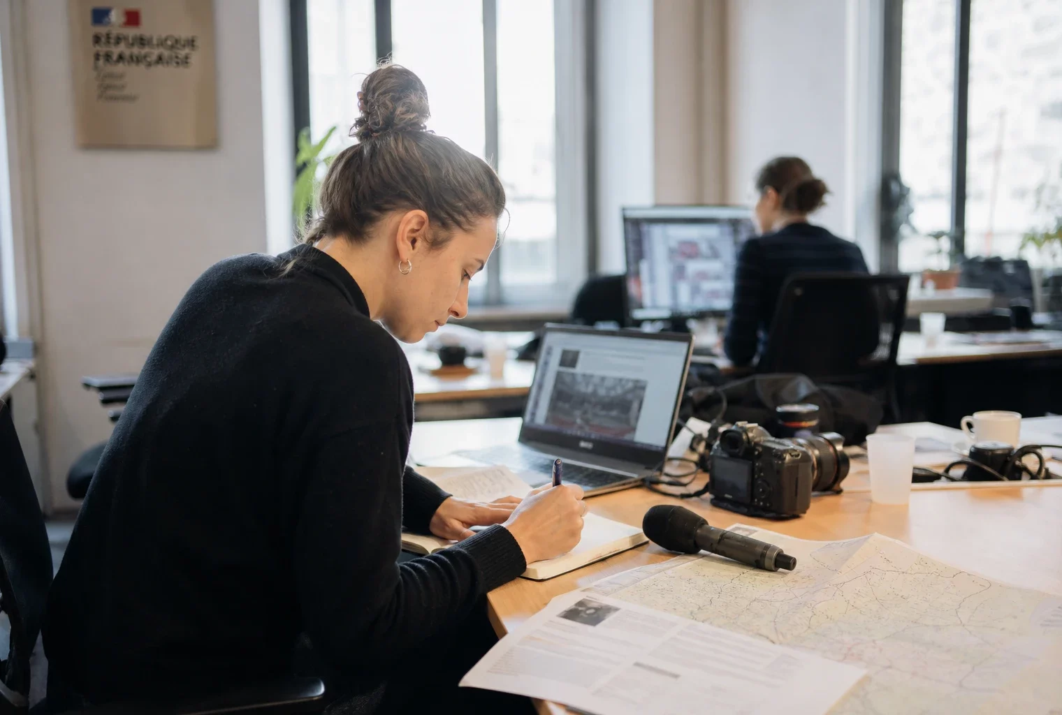 Journaliste dans une rédaction locale préparant un sujet politique, avec micro, carnet et écrans flous.