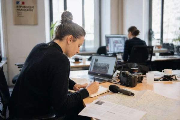 Journaliste dans une rédaction locale préparant un sujet politique, avec micro, carnet et écrans flous.