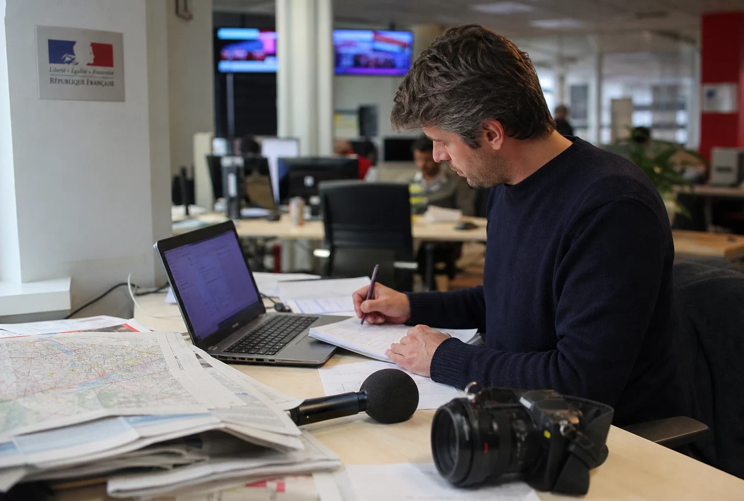 Journaliste dans une rédaction locale préparant une analyse politique avec carnet, micro sans logo et carte floue.