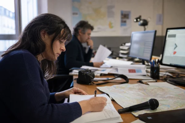 Journaliste en rédaction locale préparant un sujet sur Saint-Denis, avec carnet, carte floue et micro sans logo.