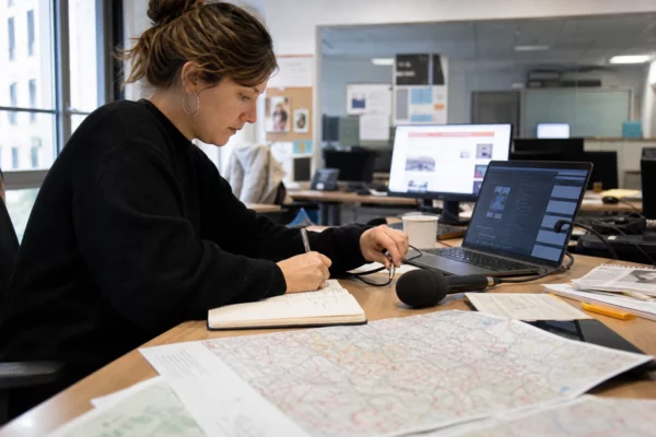 Un journaliste en rédaction prépare un sujet territorial avec carnet, carte papier floue et micro sans logo.