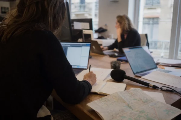 Journaliste préparant un sujet sur la fin de vie dans une rédaction française lumineuse, avec micro, carnet et écrans flous.
