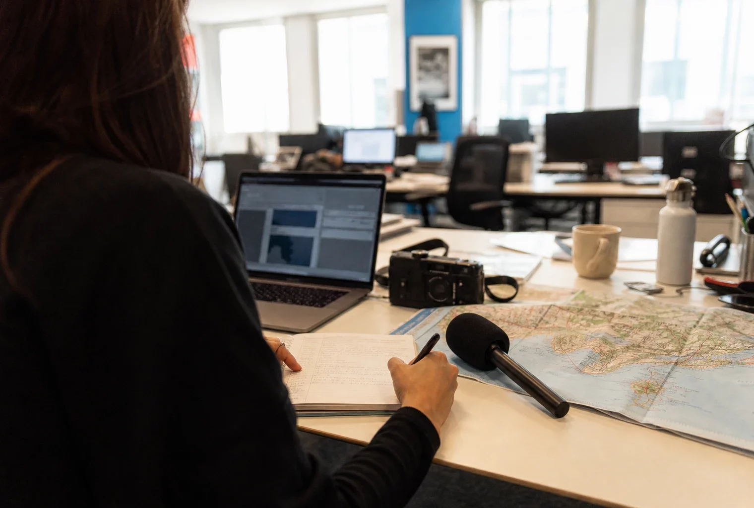 Un journaliste prépare un sujet sur la commémoration de Nice dans une rédaction française lumineuse, avec carnet, carte floue et micro.