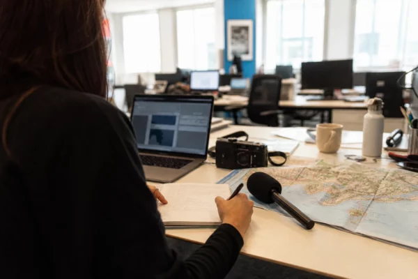 Un journaliste prépare un sujet sur la commémoration de Nice dans une rédaction française lumineuse, avec carnet, carte floue et micro.